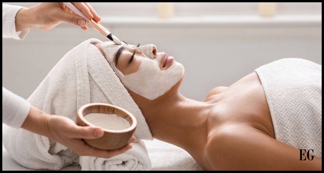 Mujer recostada con una toalla envuelta alrededor de la cabeza y el cuerpo, recibiendo un tratamiento facial. Una persona con un pincel está aplicando una mascarilla facial desde un cuenco de madera que sostiene en la otra mano. La mujer está relajada con los ojos cerrados, disfrutando del proceso de cuidado de la piel en un entorno que parece ser un spa o un salón de belleza.