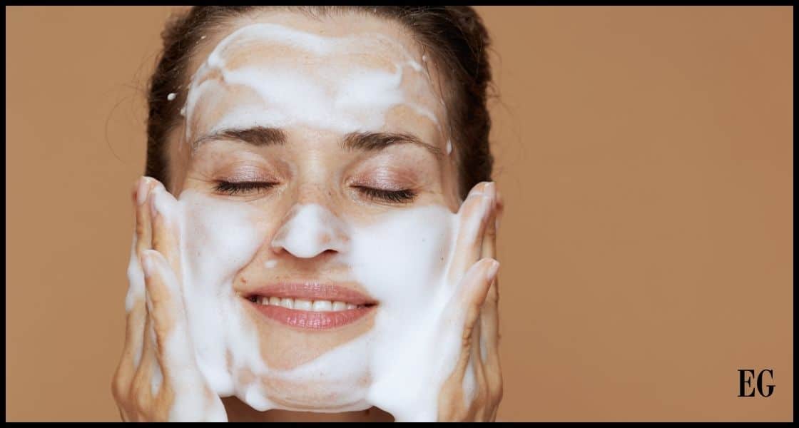 Imagen de una mujer con los ojos cerrados y una sonrisa leve, aplicándose un limpiador facial o espuma en la cara con ambas manos. Su rostro está cubierto parcialmente con el producto de limpieza, lo que indica que está en medio de una rutina de cuidado de la piel. El fondo es de color neutro